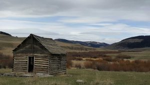 small, aged abandoned cabin sitting in the middle of a Montana field with mountains in the background