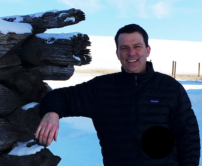 3RM owner Mike Rivers standing in a snowy field smiling while leaning on an old cabin