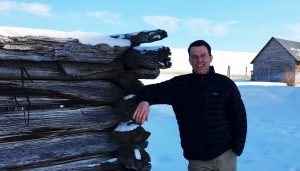 3RM owner Mike Rivers standing in a snowy field smiling while leaning on an old cabin